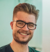 Ein weißer, blonder Mann mit stylischer Brille lächelt in die Kamera.