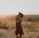 Ein Paar umarmt sich zärtlich in einer Wüstenlandschaft bei Sonnenuntergang, wobei die Frau in einem fließenden, weinroten Kleid sich in die Arme des Mannes lehnt.