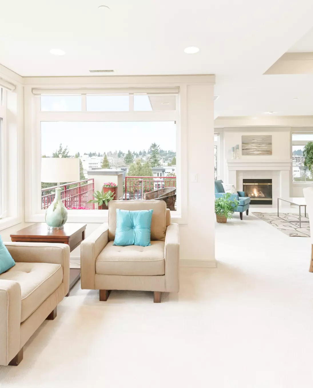 A bright and airy living room features beige armchairs with vibrant blue cushions, set against large windows overlooking a balcony
