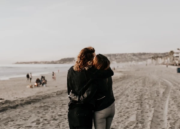 Two people embrace while walking along a sandy beach, with a few others in the distance and a calm ocean on the left under a cloudy sky.