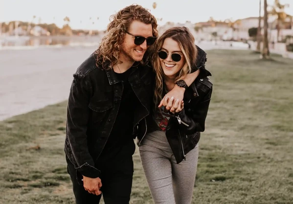 A couple wearing sunglasses and casual jackets walks closely together on a grassy area, smiling and enjoying a relaxed outdoor setting.
