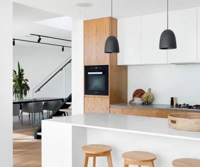 Modernioje virtuvėje yra elegantiška balta sala su dviem mediniais taburetėmis, viršuje kabančiais pakabinamais šviestuvais ir medinėmis spintelėmis. Fone matoma valgomojo zona su ilgu stalu ir juodomis kėdėmis.