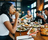 Skupina lidí si společně užívá večeři v restauraci, v popředí je žena, která si dává sousto.