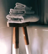 A stack of neatly folded white towels sits on a wooden stool with slender legs, set against a softly lit bathroom background.