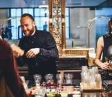 A smiling bartender in a black shirt is preparing drinks behind a counter, surrounded by various bottles and plastic cups, while a woman stands nearby with a shaker