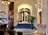 A stylish hotel lobby features elegant seating with plush chairs and a patterned rug, leading to an arched doorway framed by glass panels.