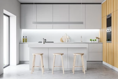 Moderní kuchyň se vyznačuje elegantním bílým ostrůvkem se třemi dřevěnými stoličkami, doplněným minimalistickými bílými skříňkami a světlou dřevěnou akcentní stěnou.