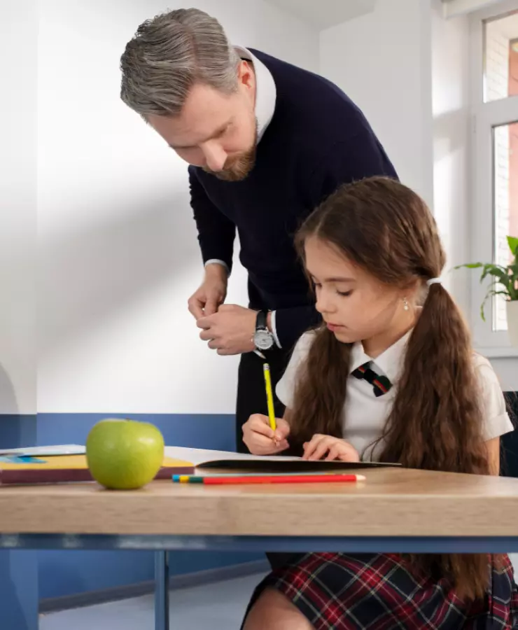 a little boy is writing in a notebook and a teacher is standing next to him