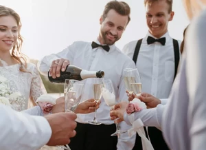 Skupina lidí oblečených ve formálním oblečení slaví, muž v bílé košili a černém motýlku nalévá šampaňské do sklenic