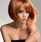 Une femme aux cheveux roux flamboyants coupés au carré fixe intensément l'objectif, vêtue d'un haut noir sans bretelles sur fond neutre.