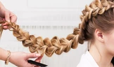Une personne réalise une tresse épaisse et complexe dans les longs cheveux blonds d'une femme. La tresse part du haut de sa tête et s'étend vers l'extérieur, dévoilant ainsi la finesse de ses motifs.
