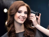 Une femme aux cheveux bruns ondulés sourit pendant qu'on lui coiffe les cheveux.