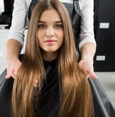 Une femme aux longs cheveux bruns et raides est assise sur un fauteuil de salon, tandis qu'une coiffeuse se tient derrière elle, tenant délicatement ses cheveux.
