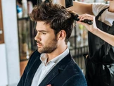 Un homme aux cheveux coiffés est assis sur un fauteuil de salon, vêtu d'un blazer foncé et d'une chemise blanche, tandis qu'un coiffeur utilise une brosse et un sèche-cheveux pour lui coiffer les cheveux.