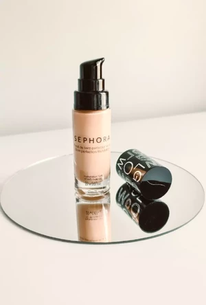 Na kulatém zrcadle stojí lahvička make-upu Sephora s pumpičkou a poblíž leží její černý uzávěr, který odráží minimalistické prostředí.