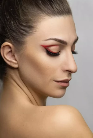 Žena s úhledně svázanými vlasy a výraznou červenou oční linkou hledí dolů a na neutrálním pozadí zdůrazňuje svůj elegantní profil.