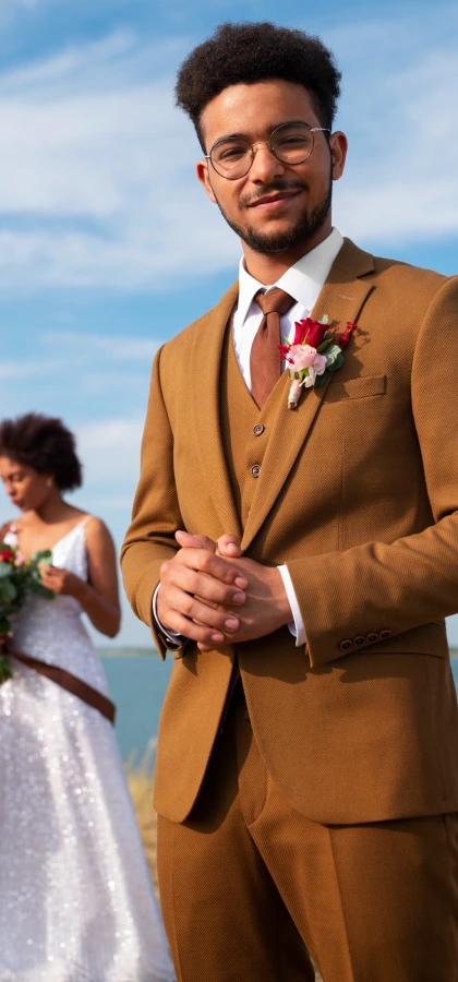 Muškarac u smeđem odijelu i naočalama stoji vani, smiješi se sklopljenih ruku i nosi boutonniere.