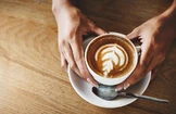 Eine Person hält eine weiße Tasse Kaffee mit kunstvoller Latte-Art auf einem Holztisch.