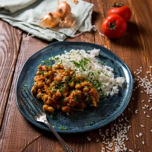 A rustic wooden table with a plate of chickpea curry and rice, garnished with herbs, accompanied by a tomato and bread rolls