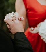Ein Paar hält zärtlich Händchen, die Frau trägt ein auffälliges rotes Kleid und einen Ring mit Schmetterlingsmotiv.