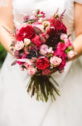 Eine Person in einem weißen Kleid hält einen farbenprächtigen Brautstrauß, der aus einer Mischung roter, rosa und violetter Blumen, darunter Rosen und Dahlien, sowie grünen Blattwerkakzenten besteht.