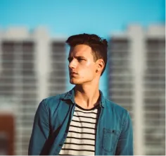 a young guy in a denim shirt and a striped T-shirt looks into the distance against the backdrop of high-rise buildings