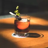 Auf einem Holztisch steht ein Glas mit dunkelbernsteinfarbenem Cocktail, garniert mit einem Spieß, an dem eine Kirsche steckt.