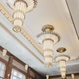 Elegante Art-Deco-Kronleuchter mit aufwendigen geometrischen Mustern hängen von der hohen Decke eines geräumigen, hellen Raumes mit großen Fenstern und klassischer Holzvertäfelung.