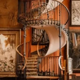 Eine Wendeltreppe mit kunstvollen schmiedeeisernen Geländern führt in einen warm beleuchteten Raum hinauf, der mit großen gerahmten Kunstwerken gefüllt ist.