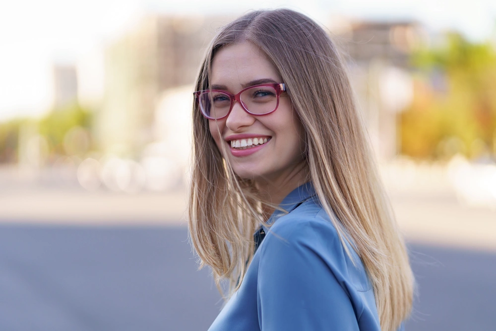ragazza bionda che indossa occhiali con montatura rossa e una giacca blu sorride ampiamente