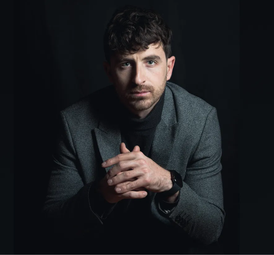 Man in dark suit with clasped hands against black background
