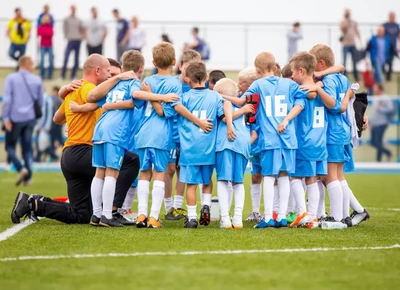 A youth soccer team in blue uniforms huddles together on a grassy field, with their coach kneeling beside them for a pep talk.