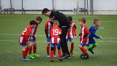 A group of young children in red and white striped soccer uniforms huddles around a coach on a grassy field, listening intently. A child in a blue goalkeeper outfit stands nearby.