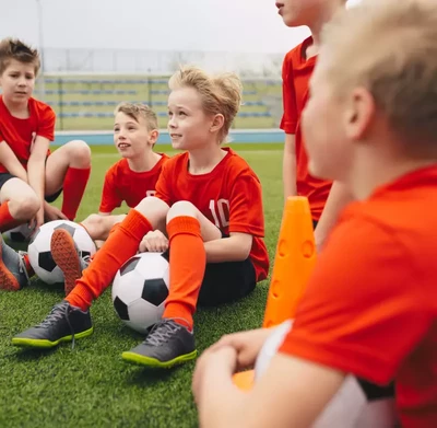 A group of young boys in red soccer uniforms sit on a grassy field, each with a soccer ball.