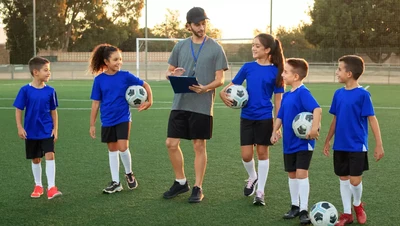 A group of five children in blue jerseys stand on a soccer field, each holding a soccer ball, while a coach in a cap and shorts reviews notes on a clipboard.