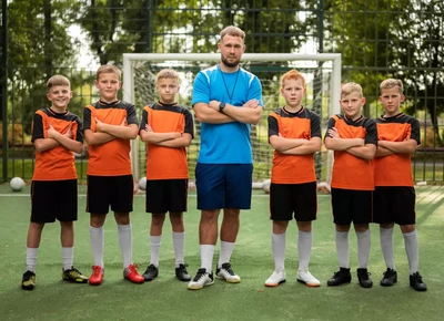 A soccer coach stands with six young players in orange and black uniforms, all posing confidently with crossed arms on a field in front of a goal.