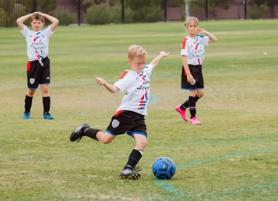 Children in colorful soccer uniforms, one in black with neon green socks and another in red, energetically chase a soccer ball on a vibrant green field.