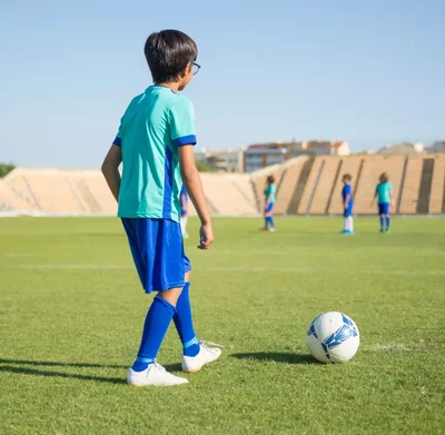 A young boy in a teal and blue soccer uniform stands on a grassy field, poised to kick a soccer ball.