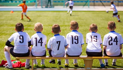 Six young soccer players wearing white jerseys with numbers sit on a bench, watching a game on a grassy field.