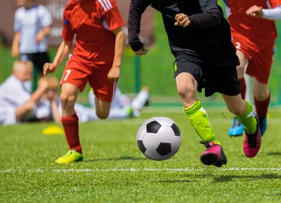 Young soccer players in action on a vibrant green field, with one in a black uniform and neon green socks dribbling a black-and-white ball, pursued by opponents in red uniforms.