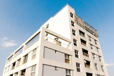 A modern, white apartment building with multiple balconies is set against a clear blue sky, showcasing its clean lines and large windows
