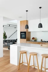 A modern kitchen features a sleek white island with three wooden stools, pendant lights hanging above, and light wood cabinetry.