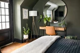 A stylish bedroom features a large round mirror above a wooden desk with a brown chair, flanked by potted plants and a tall floor lamp
