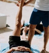 Einer Person, die im Freien auf einer Matte liegt, wird von einer neben ihr stehenden Person, die blaue Shorts und ein weißes Hemd trägt, das Bein gedehnt.