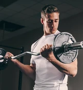 Ein Mann in einem weißen Sportshirt führt in einem Fitnessstudio Bizepscurls mit einer Langhantel durch und konzentriert sich dabei intensiv auf seine Übung.