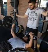 Ein Mann stemmt beim Bankdrücken eine schwere Langhantel mit je 20 kg Gewichten auf jeder Seite, während ein anderer Mann in einem weißen T-Shirt ihn dabei beobachtet.