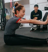Eine Frau in einem gestreiften Shirt und schwarzen Leggings sitzt auf dem Boden einer Turnhalle und streckt die Arme nach vorn.