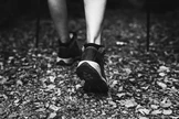 Eine Person in Sportschuhen geht auf einem laubbedeckten Waldweg; Wanderstöcke sind sichtbar, was auf eine Wanderung durch eine natürliche Umgebung hindeutet.
