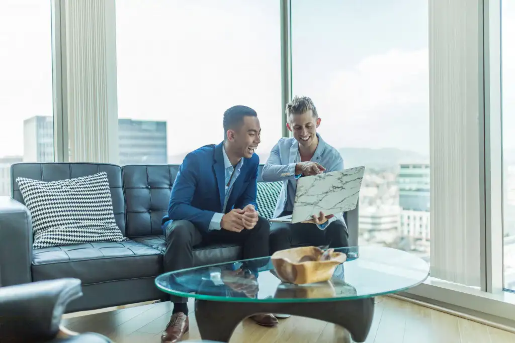 2 men are sitting on the couch and looking at a laptop