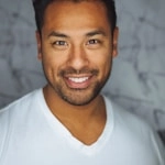 A smiling man in a white V-neck shirt stands against a soft-focus marble-patterned background.